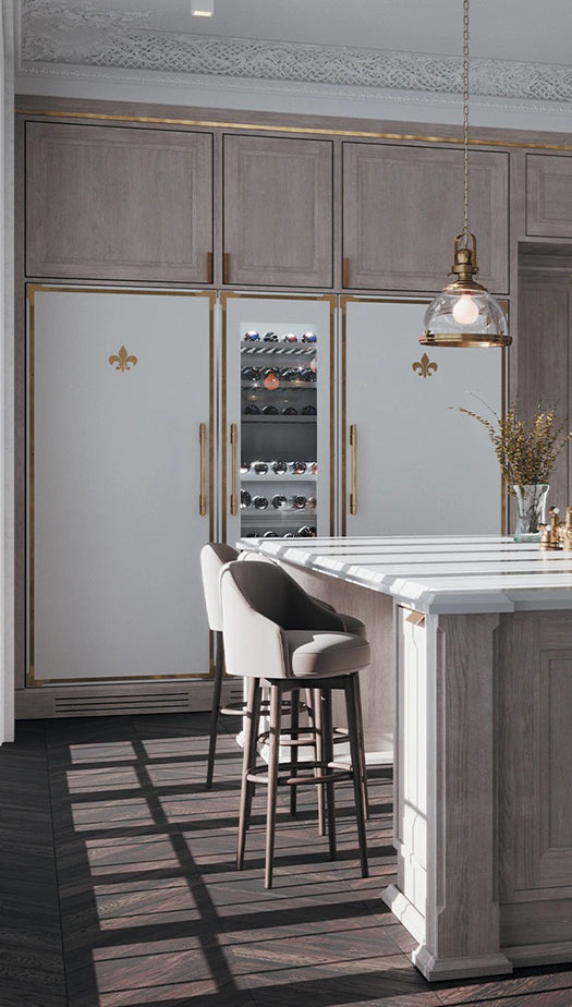 A luxurious kitchen features integrated, paneled appliances in white with gold trim and fleur-de-lis emblems, including a central wine refrigerator and two refrigerators. The space has light gray wood cabinetry, a dark wood floor, a light-colored kitchen island with a velvet bar stool, and is lit by a single industrial-style glass pendant light.
