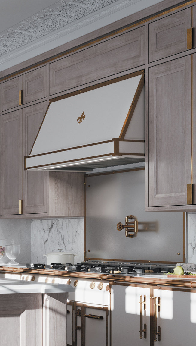 A striking close-up of a white and bronze range hood with a gold fleur-de-lis emblem, mounted above a professional cooktop and a white marble backsplash. The kitchen features light gray wood cabinetry with gold bar pulls, a bronze pot filler faucet on a metal panel, and a white and bronze range visible below.