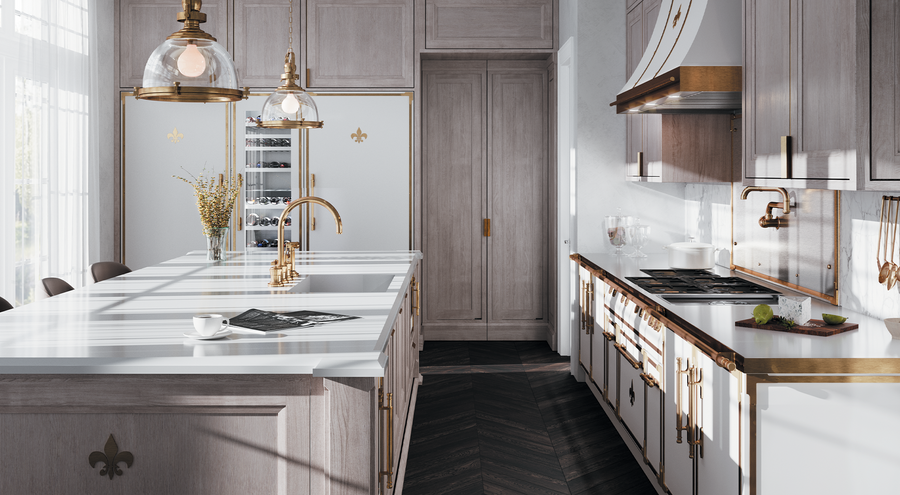 A bright, French-inspired luxury kitchen featuring a large light gray wood island with a white marble countertop, a gold faucet, and two industrial-style pendant lights. The kitchen includes a white and bronze range and hood, light gray cabinetry with gold hardware, a dark herringbone wood floor, and paneled refrigerators with gold fleur-de-lis emblems.
