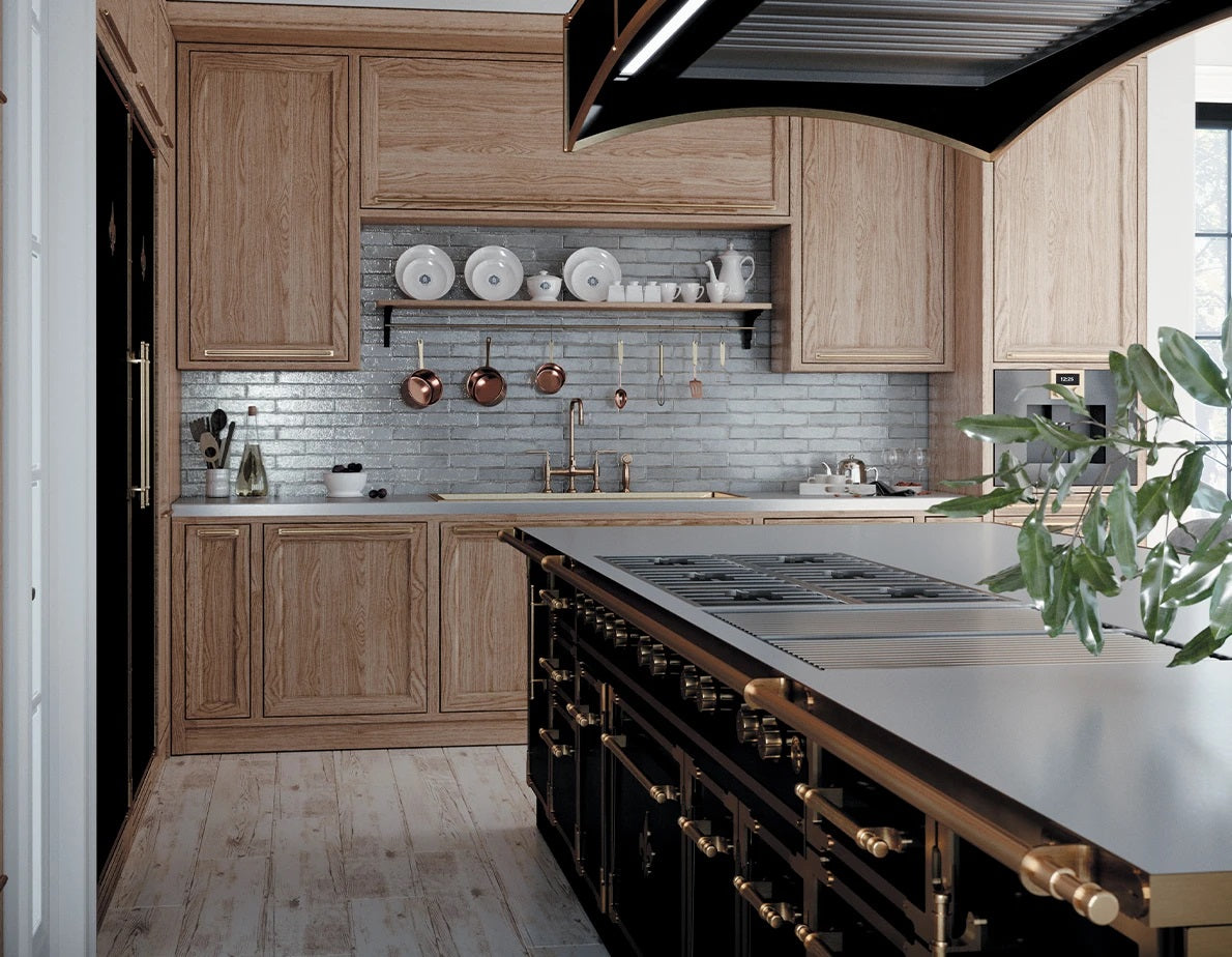 A striking, high-end kitchen featuring a large black professional range island with a metal countertop and brass handles. The surrounding walls have natural wood cabinetry, a blue-gray subway tile backsplash, a pot filler faucet, and open shelving with copper pots and white dishware.