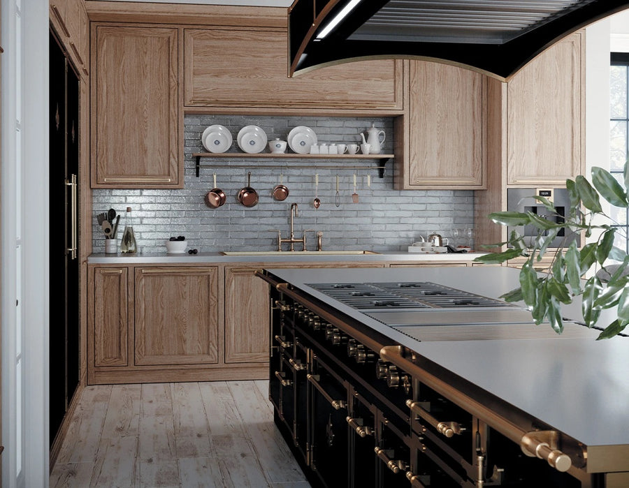 A striking, high-end kitchen featuring a large black professional range island with a metal countertop and brass handles. The surrounding walls have natural wood cabinetry, a blue-gray subway tile backsplash, a pot filler faucet, and open shelving with copper pots and white dishware.