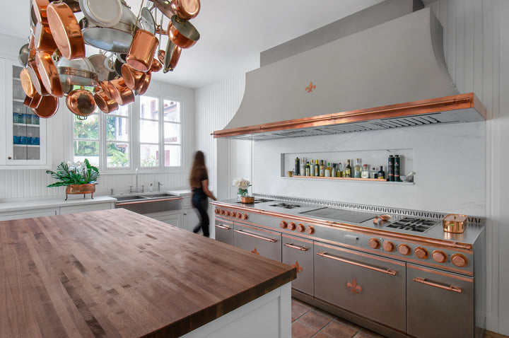 Silver Kitchen Range With Rose Gold Handles and Burners and Silver Exhaust Hood above it. Copperware hanging above wooden tabletop