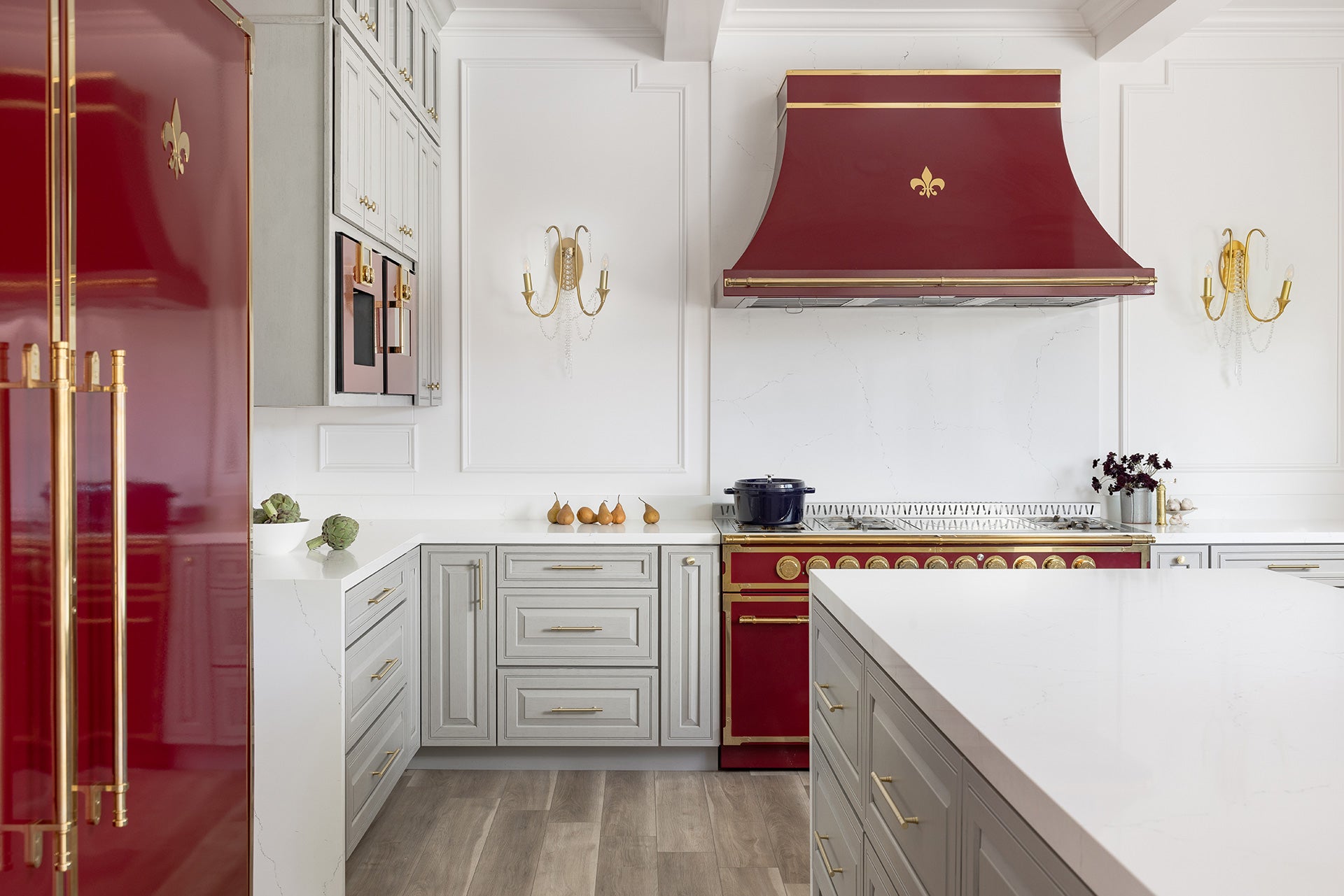 A luxurious French-style kitchen with a maroon and gold range and a matching range hood and refrigerator, both featuring a fleur-de-lis emblem. The kitchen has light gray cabinetry with gold hardware, white countertops and backsplash, and ornate wall sconces flanking the hood.