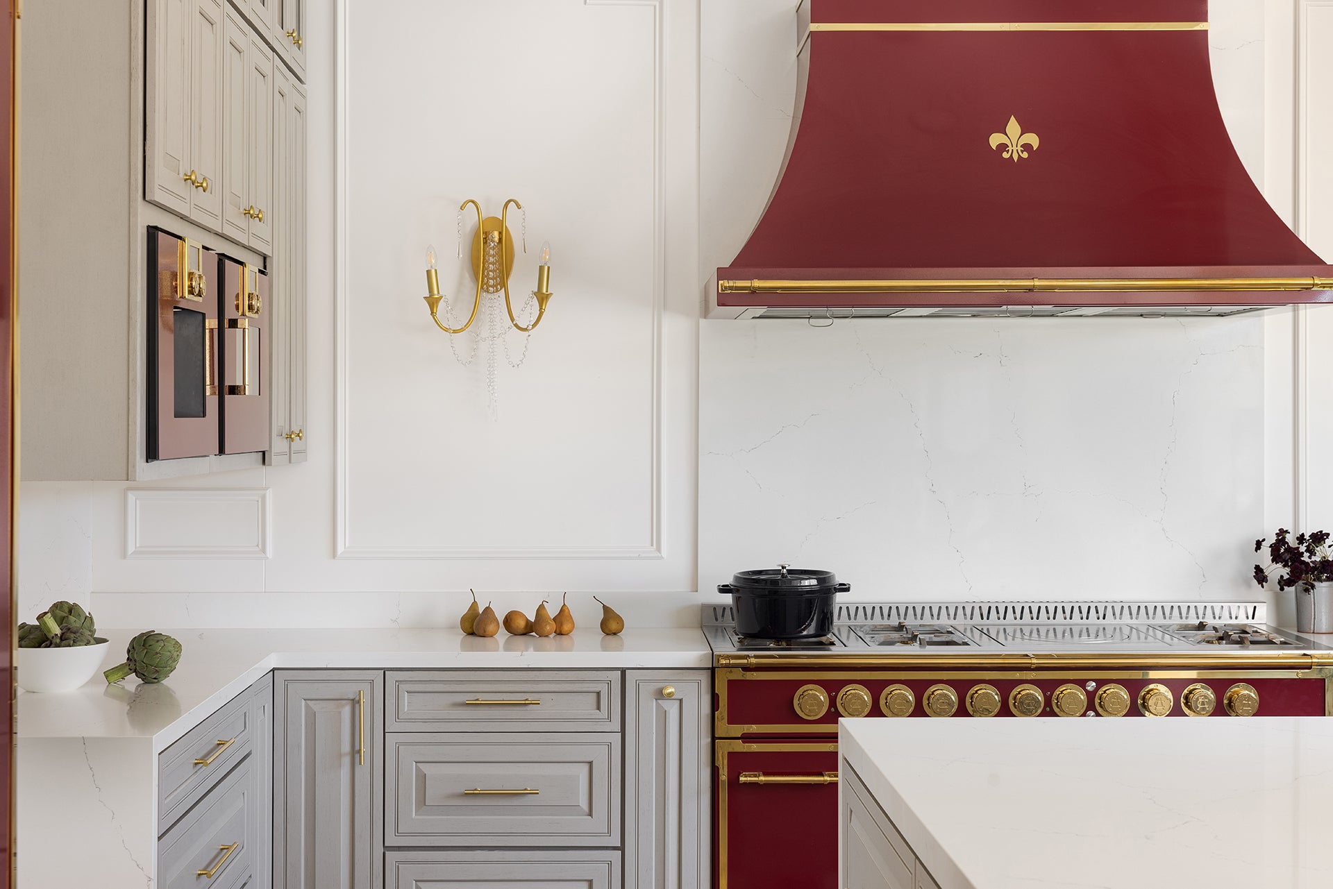 A high-end kitchen featuring a vibrant maroon and brass range with a matching custom range hood adorned with a fleur-de-lis. The space boasts elegant light gray cabinets, white marble countertops, a clean white paneled wall with a decorative gold sconce, and subtle details like pears on the counter.
