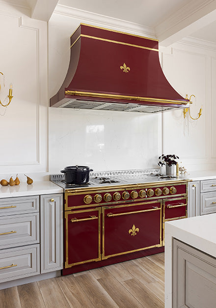 A striking image of a maroon and brass professional range and its matching custom range hood, both featuring a fleur-de-lis emblem, as the centerpiece of a luxury French-style kitchen. The range is set between light gray cabinets with gold hardware, a white marble backsplash, and surrounded by ornate white wall paneling.