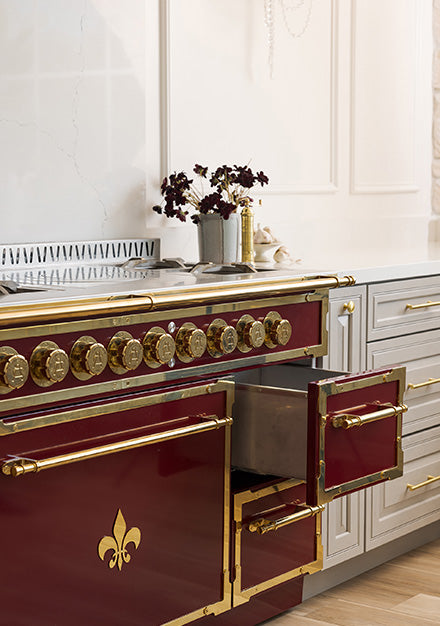 A close-up of a luxurious, French-style range in maroon and polished brass, featuring a fleur-de-lis emblem on the lower panel and detailed burner controls. The image also shows a small storage drawer pulled open, alongside light gray cabinets and a white marble backsplash.