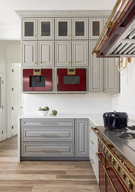 A French-style luxury kitchen featuring light gray cabinetry with gold hardware, a white countertop and backsplash, and wood-look flooring. The design includes stacked upper cabinets and a pair of built-in maroon appliances (possibly a coffee maker and microwave) integrated into the wall.