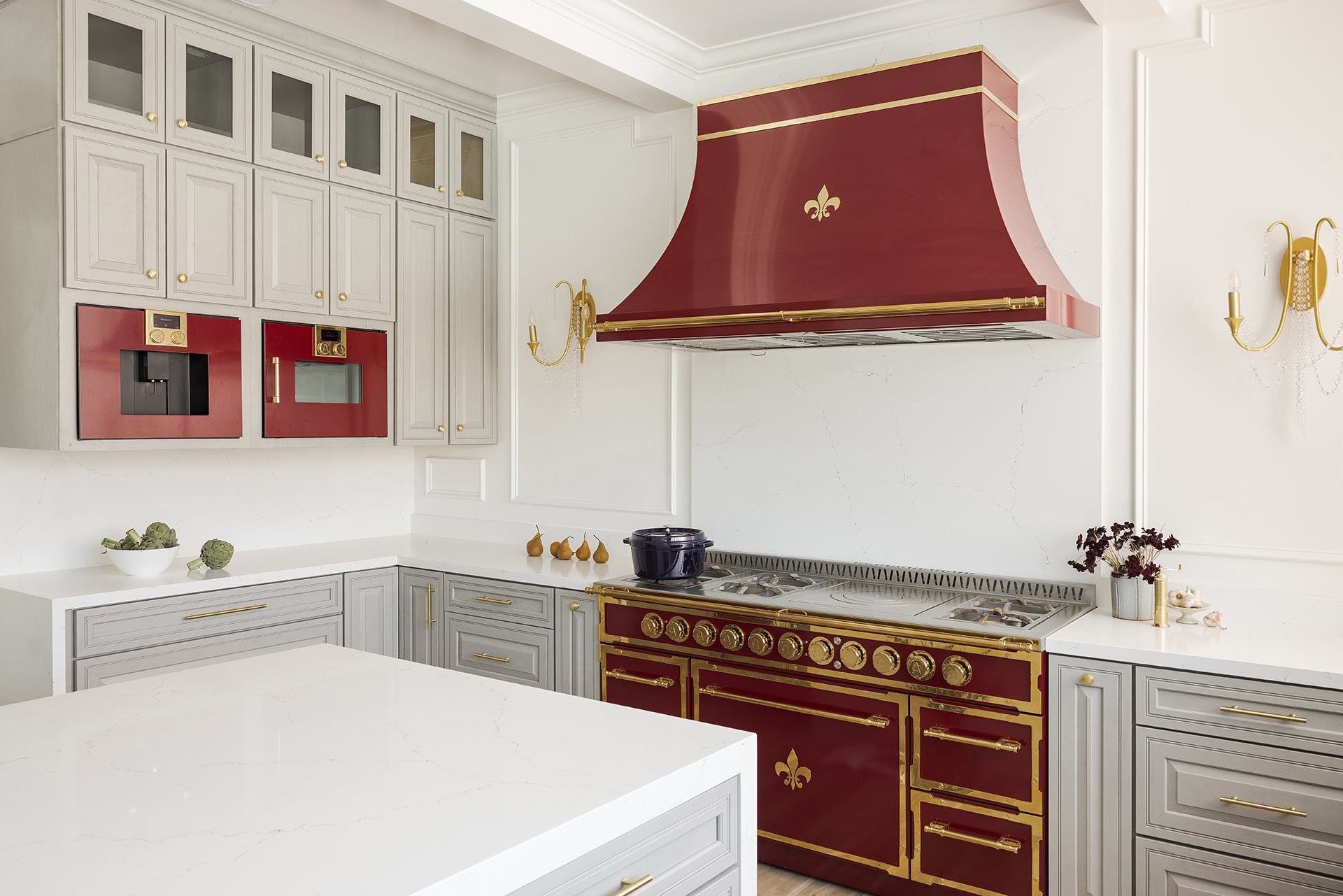 A bright and luxurious French-style kitchen featuring a stunning maroon and brass professional range and a custom range hood with a fleur-de-lis emblem. The room has light gray paneled cabinetry with gold hardware, white marble countertops and backsplash, and a set of matching built-in maroon appliances on the left wall.