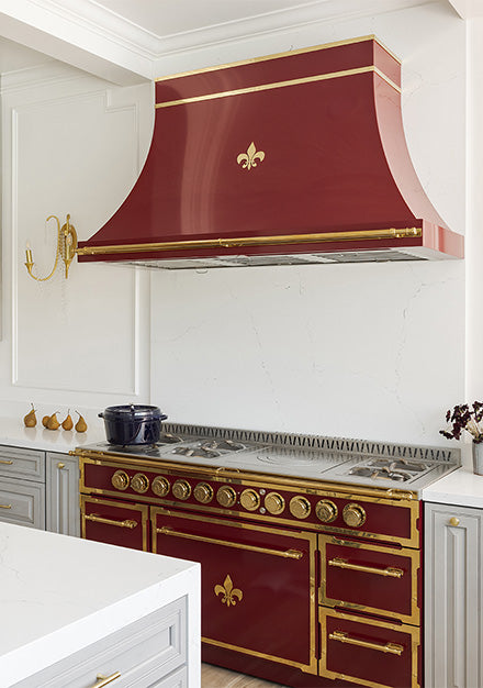 A close-up of a luxury French kitchen highlighting a maroon and brass professional range and a matching curved range hood, both decorated with a fleur-de-lis motif. The range is set against a white marble backsplash and flanked by light gray cabinetry with white quartz countertops.