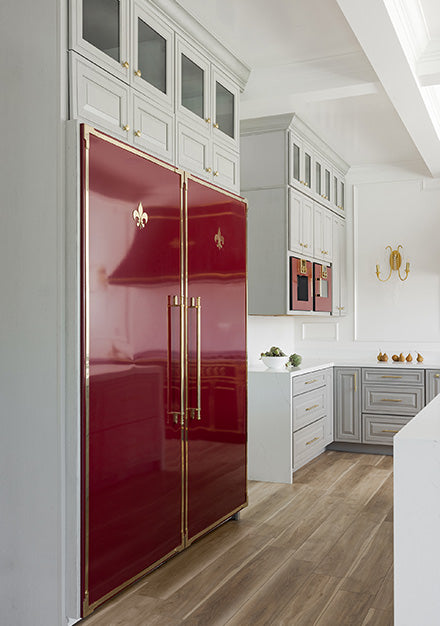 A stunning French-style kitchen featuring a large built-in double refrigerator in a glossy maroon finish with polished brass trim and handles, topped with a fleur-de-lis emblem. The kitchen has light gray cabinetry with gold hardware, white countertops, and wood-look flooring.