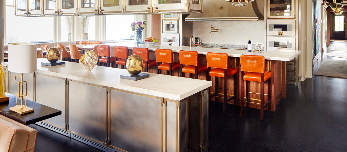 Luxury open-concept kitchen and bar featuring a long white island with vibrant orange leather bar stools, metallic cabinetry, and integrated wall ovens