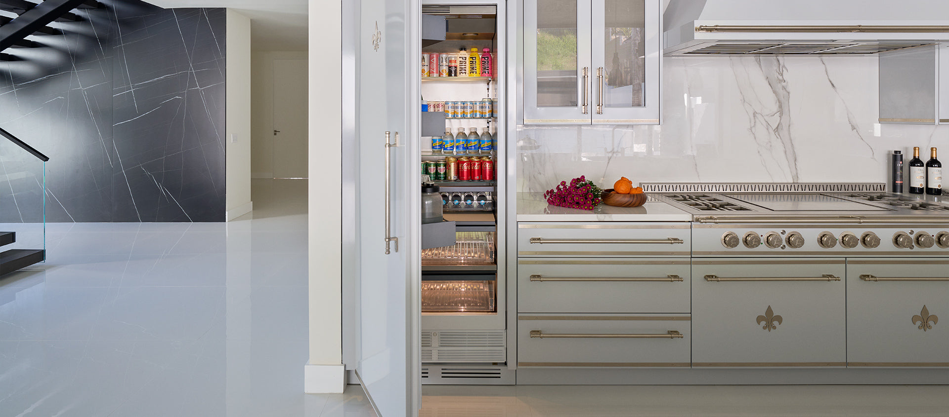 A high-end kitchen with a built-in, stainless steel refrigerator that has its door open, revealing a full stock of drinks and a glass-front freezer drawer below.