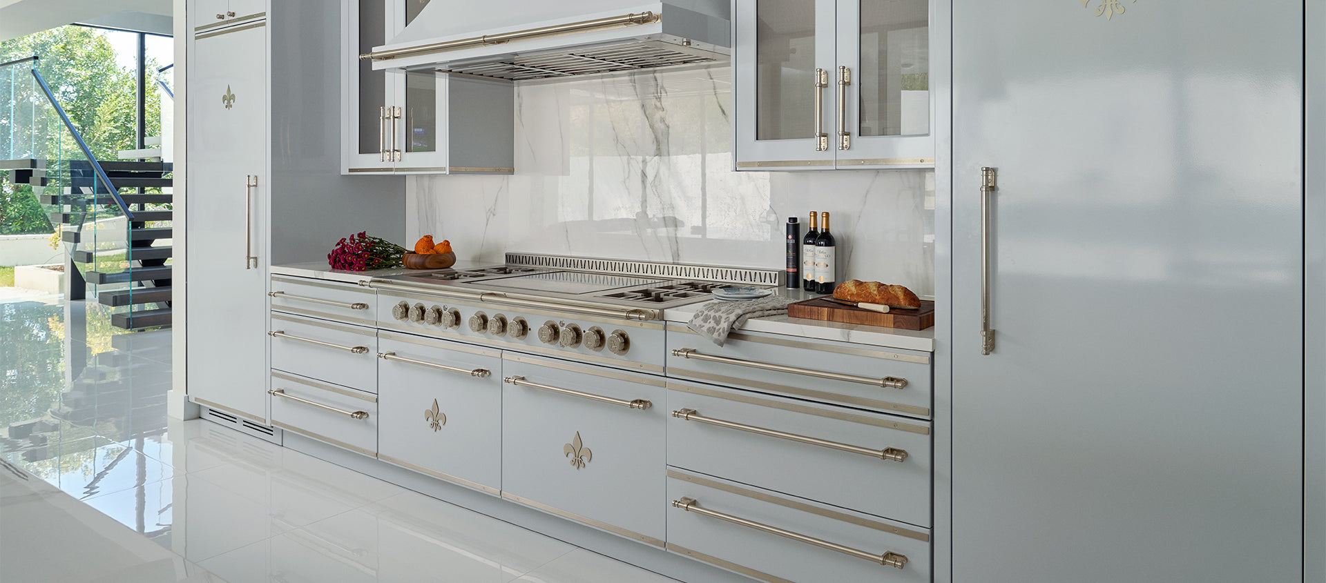 A luxury kitchen with light gray cabinetry, gold hardware featuring a fleur-de-lis emblem, a large professional range with multiple burners, and a built-in refrigerator, all under a matching range hood.